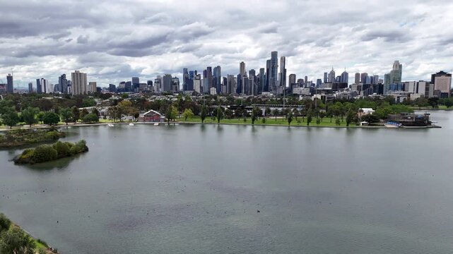 Smooth cinematic drone pan across Albert Park Lake, revealing the Melbourne skyline and modern urban architecture under dramatic cloudy skies.