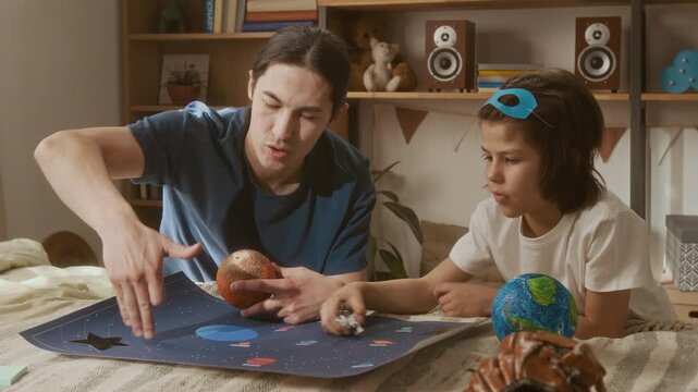 Asian young man teaching school-age multiethnic boy about astronomy using planet models and space poster on bed. Ideal for promoting homeschooling, STEM education, and e-learning platforms