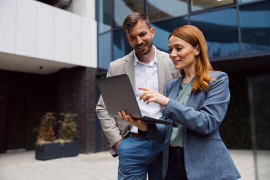 Two diverse business professionals standing outside a modern glass office building, looking at a laptop screen and discussing a project during a meeting.