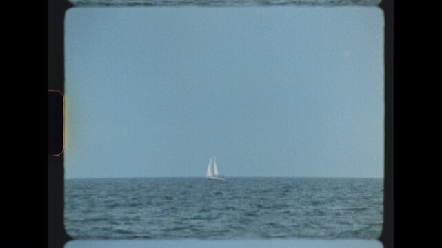 A single white sailboat glides far out on a calm blue sea beneath a soft hazy sky on a quiet summer day captured on retro super 8 film with gentle grain