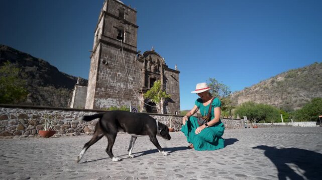 A senior woman pets and greets a stray dog at San Javier Mission near Loreto in Baja California Sur at sunrise