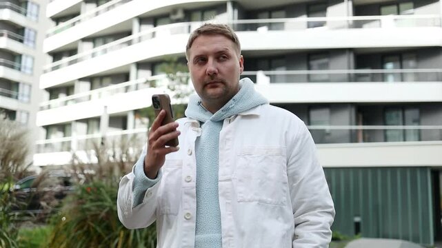 Thoughtful young caucasian man wearing a smart casual outfit with a tie, holding a mobile phone and texting a message while standing outside a modern residential or office building complex