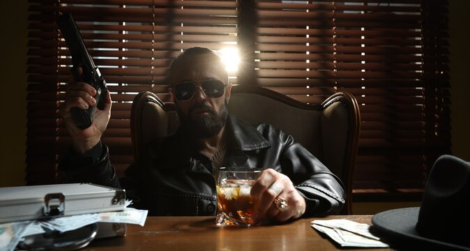 Mafia. Man with gun and whiskey at table in dark room