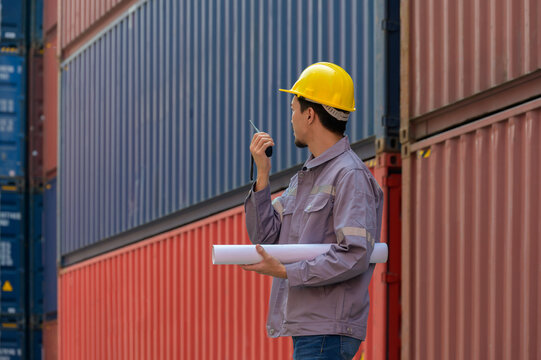 Asian male logistics coordinator or industrial engineer wearing yellow safety hardhat holding blueprints and talking on walkie talkie at  shipping container terminal