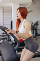Fototapeta premium Red haired woman exercising on elliptical machine in gym, working out for fitness and healthy lifestyle