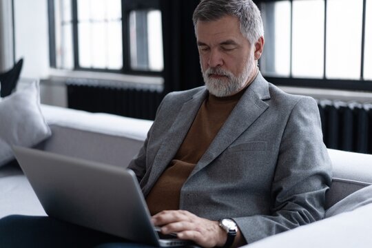Happy smiling mature man wearing suit looking at laptop using computer watching webinar sitting at sofa.