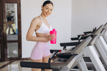 Fototapeta premium Sporty young woman in gym wearing sportswear, standing on treadmill with shaker, practicing fitness for health