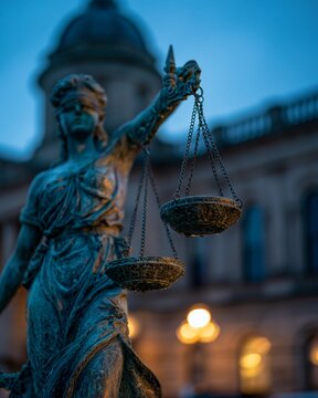 Closeup of weathered bronze Lady Justice statue with rainsoaked surface and a tilted scale set against a softly blurred historic courthouse illuminated by moody bluehour light