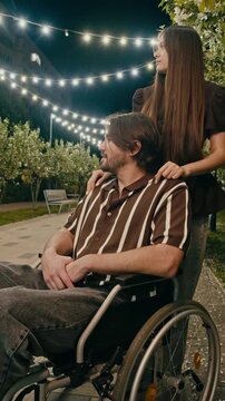 Young wheelchair user speaks with woman standing beside him in illuminated garden at night. Warm string lights and flowering trees create intimate accessible lifestyle scene about care and connection.