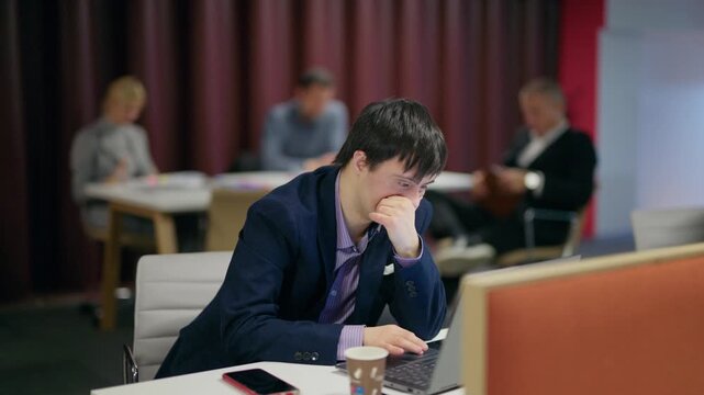 A businessperson with Down syndrome intently uses a laptop in a collaborative workspace. An employee focused on his computer in a professional office environment
