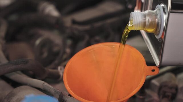 Close-up view of motor oil being poured from a container into an orange funnel inside an engine compartment, with blurred mechanical parts in the background