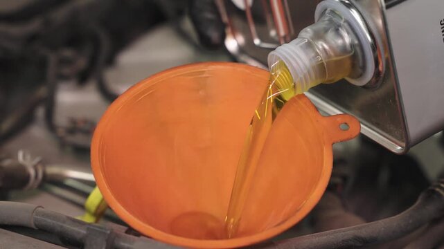 Close-up of golden liquid flowing from a metal container into an orange plastic funnel in an industrial workshop, with blurred machinery, hoses, and soft