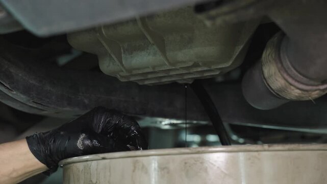 Gloved hand working beneath the engine area of a vehicle in a dim workshop, catching a dark fluid drip into a container during inspection and maintenance