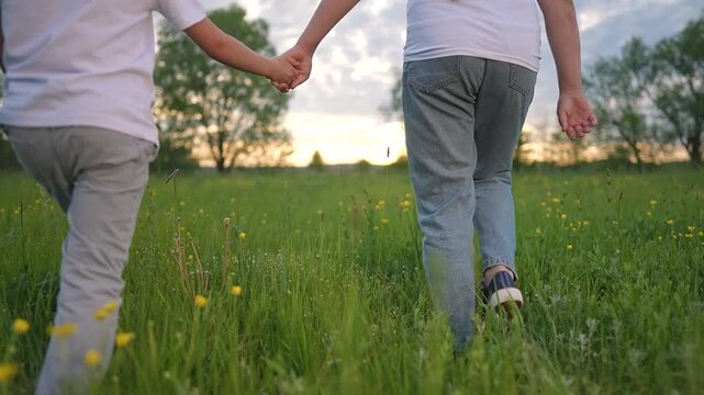 Children holding hands walk in field. Girl and boy surrounded by tall grass. Nature frames their movement. Together they cross green space. Grass brushes children legs. Girl steps boy through grass