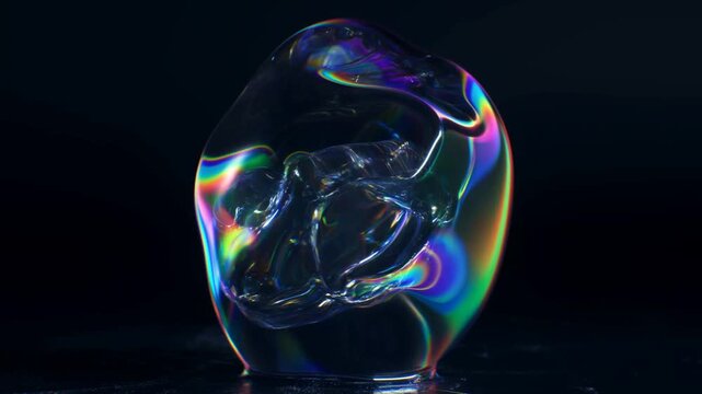 Static shot of iridescent glass sculpture rotating on reflective tabletop in dark studio, close up view of transparent orb turning and tilting