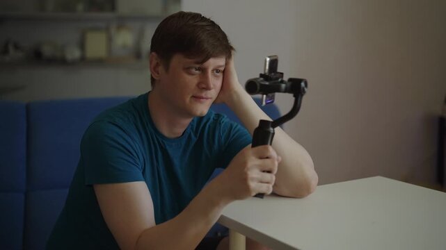 Young man sitting at table filming himself on mobile phone mounted on gimbal stabilizer for vlog, leaning head on hand talking during live broadcast session at home. Shooting in slow motion