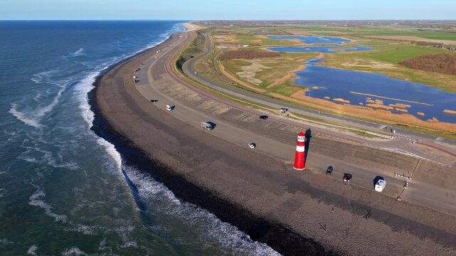 Aerial drone panoramic view of a coastal road on a stone dike with a red and white lighthouse, parked cars, sea waves, and a marsh landscape under a blue sky in the Netherlands.