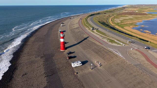 Aerial drone panorama of a scenic coastal landscape featuring a red and white lighthouse on a wide stone sea dike, with cars, a camper van, a curving highway, and rural wetlands in the Netherlands.