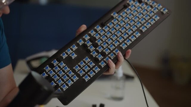 Close-up of female hands holding disassembled mechanical keyboard and using hand air blower to clean dust from blue switches, demonstrating proper peripheral maintenance