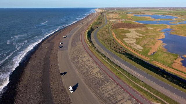 Aerial drone panorama of a long coastal sea dike with a winding highway, parked cars, rolling ocean waves, and a scenic marsh landscape stretching along the shoreline under a clear blue sky.