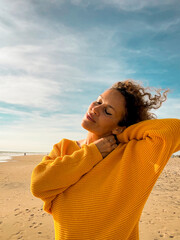 A relaxed woman in a yellow sweater stands on a sandy beach with her eyes closed, enjoying the ocean breeze and warm sunlight, curly hair blowing, portrait of calm, freedom, mindfulness, autumn  © simona