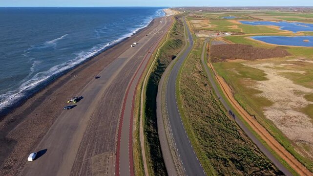 Aerial drone view of a coastal sea dike alongside rolling ocean waves, featuring a winding highway, a red bicycle path, parked vehicles, and a scenic marsh landscape stretching toward the horizon.