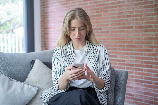 Young woman sitting on a sofa in a modern living room, focused on her smartphone as she scrolls and types, relaxed casual style with striped shirt and neutral home decor background.