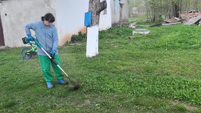 A woman trims grass in a garden using an electric trimmer. She stands near a house. The garden has green grass and trees during a sunny spring day.