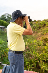Fototapeta premium Man Using Binoculars on Hilltop: An elderly man wearing a hat and yellow shirt stands on a hilltop, looking through binoculars at the lush green landscape below.