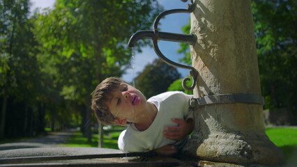 Fototapeta premium Boy leaning to drink from old stone fountain in park, enjoying fresh water outdoors, carefree childhood moment surrounded by green trees and summer light