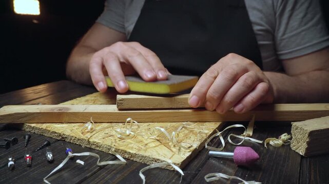 Hands of a male carpenter sanding a wooden block on a workbench with shavings and tools scattered around in a dimly lit workshop