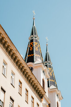 Upward view of two ornate church towers featuring steep spires topped with crosses and decorative patterned tile roofs. The pale stone facade of the adjacent