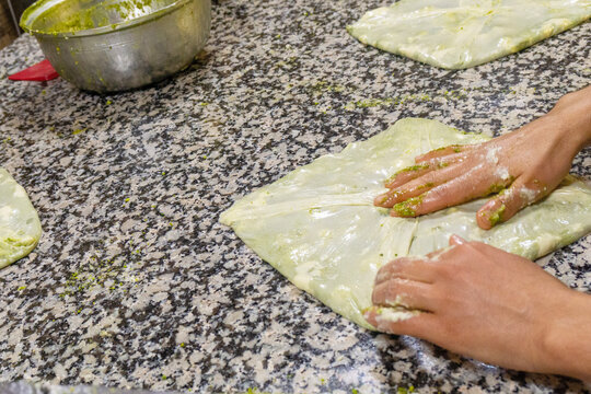 Chef spreading crushed green pistachios and fresh clotted cream on thin dough to prepare traditional Antep Katmeri pastry, close up view.