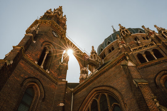 A dramatic low-angle view of an ornate brick cathedral showing tall clock tower and domed roof details against a pale sky. Sunlight creates a starburst flare