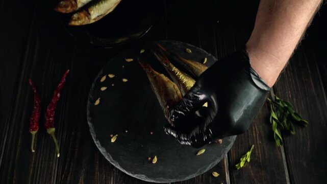 A hand in a black glove places smoked fish from a round plate onto a serving platter, on a dark wooden table, creating a rich culinary scene with chili peppers