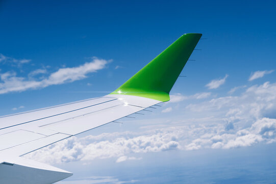View from a passenger window showing an aircraft wing and bright green winglet above a sea of white clouds under a vivid blue sky. Smooth metallic wing