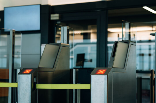 A row of stainless steel automated turnstile gates with glass panels at a transit entrance. Two gate terminals in the foreground display illuminated red X