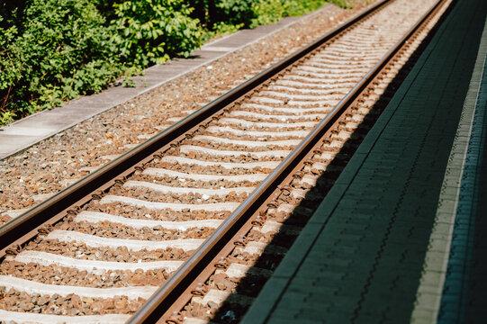 Close-up view of twin railway tracks running alongside a concrete station platform. Sunlight casts sharp shadows across the metal rails, concrete sleepers