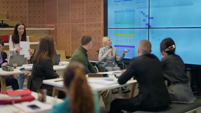 A female businessperson in a wheelchair delivers a presentation to colleagues in a modern office. Coworkers gather to discuss project timelines displayed on a large screen
