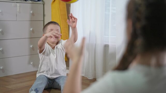 Kid laughs reaching arms to catch bright ball. Friend throws toy during indoor play. Joyful child and student share funny moment together. Kid keeps focus as they play and throw in cozy room.
