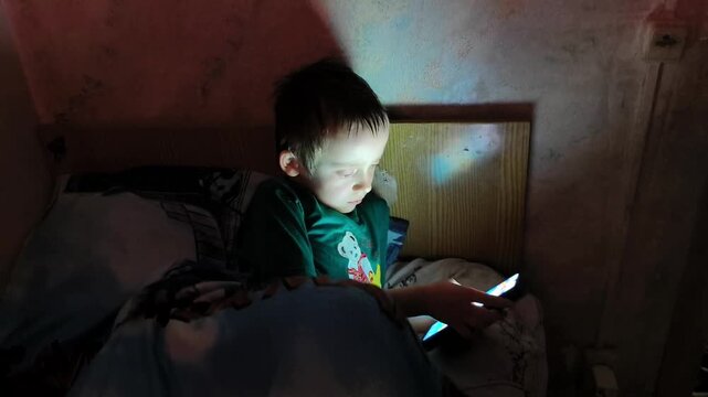 A child sits in bed with a smartphone in hand. The room is dark, and the light from the phone illuminates the child's face as they focus on the screen.