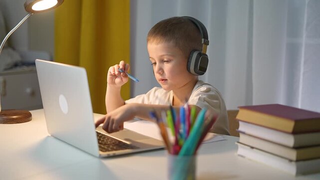 Child listens in headphones and writes homework while looking at laptop. Focused student continues homework task. Laptop connects student to digital study platform. Child handles homework alone.