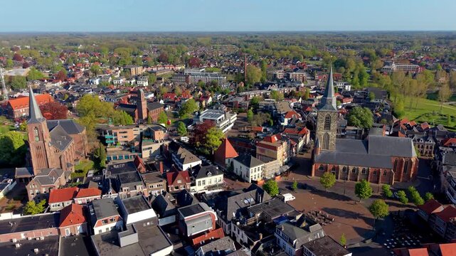 An aerial drone view of a historic European town center with two prominent brick churches, traditional houses, a public square, and lush green trees under a clear blue sky.
