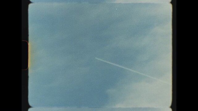 A high altitude jet aircraft flying from right to left across a soft blue sky leaving a thin white vapor trail, filmed on retro super 8 film