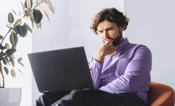 A man is seated in a chair while looking intently at a laptop. He has a thoughtful expression on his face. Sunlight streams through a window, highlighting a plant nearby