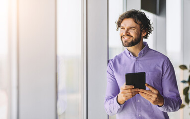 Fototapeta premium A man stands by a large window in a bright office space. He is smiling and looking outside while holding a tablet. Sunlight shines through the window creating a warm atmosphere