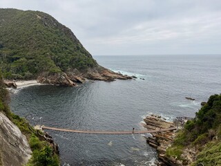 Obraz premium Suspension bridge over a scenic coastal bay in Tsitsikamma National Park in South Africa, with rugged cliffs and lush vegetation.