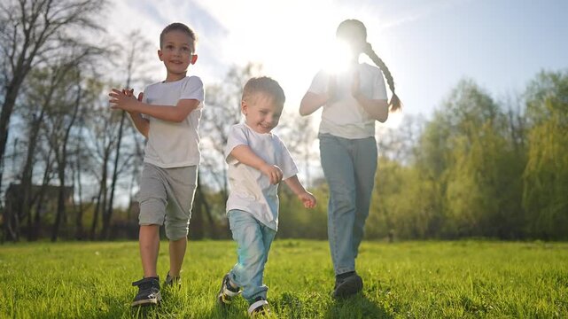 Boy runs forward on green grass. Child laughs while running in sunlight. Girl and boy follow behind smiling. Happy kids enjoy outdoor game on sunny day. Child leads way in joyful nature play.