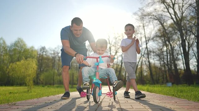 Child pedals small bike in bright park light as father leans to guide motion. Boy learns new skill while older brother claps nearby. Family support during first ride. Outdoor bike ride and learning.