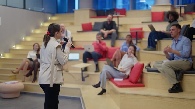 A female speaker addresses a diverse group of professionals in a modern amphitheater setting. Associates attentively listen to the presentation during a business meeting or workshop